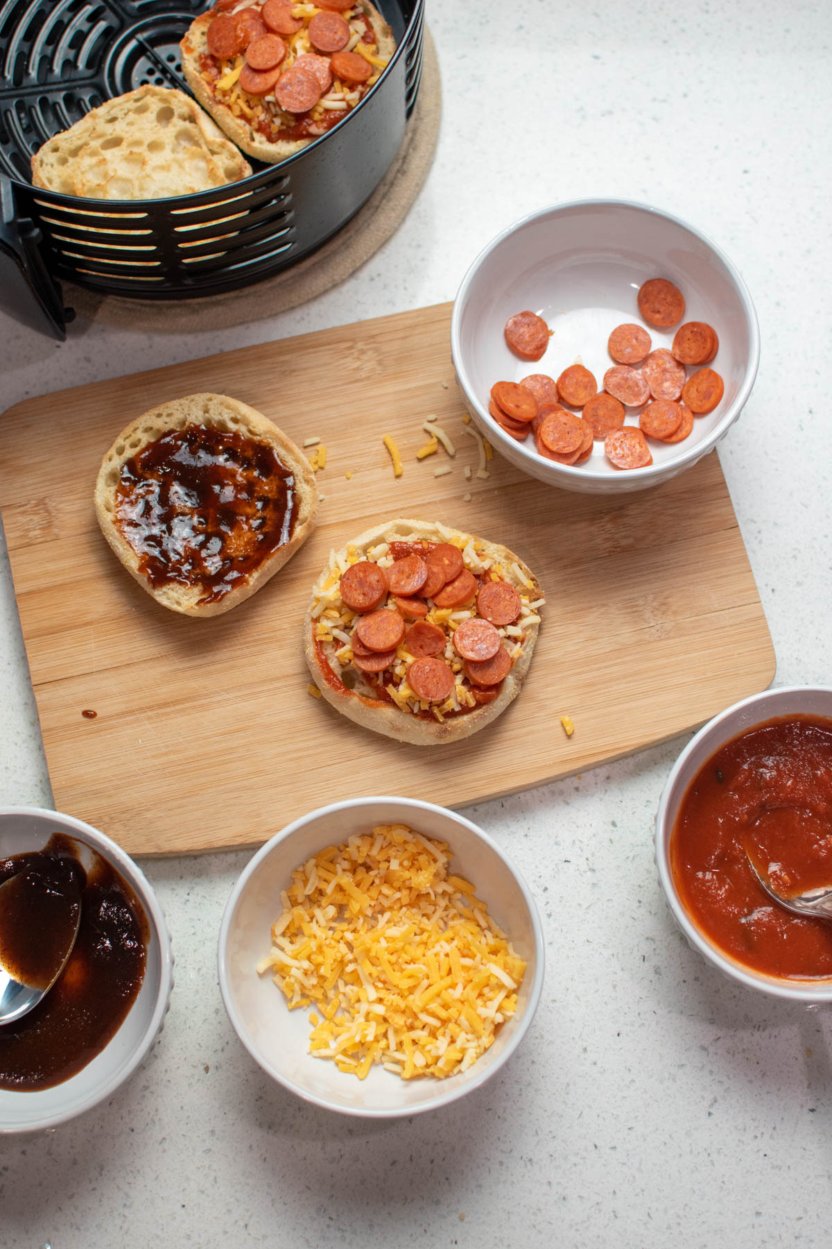 Bowls of pizza toppings on counter next to cutting board with mini pizzas on it.