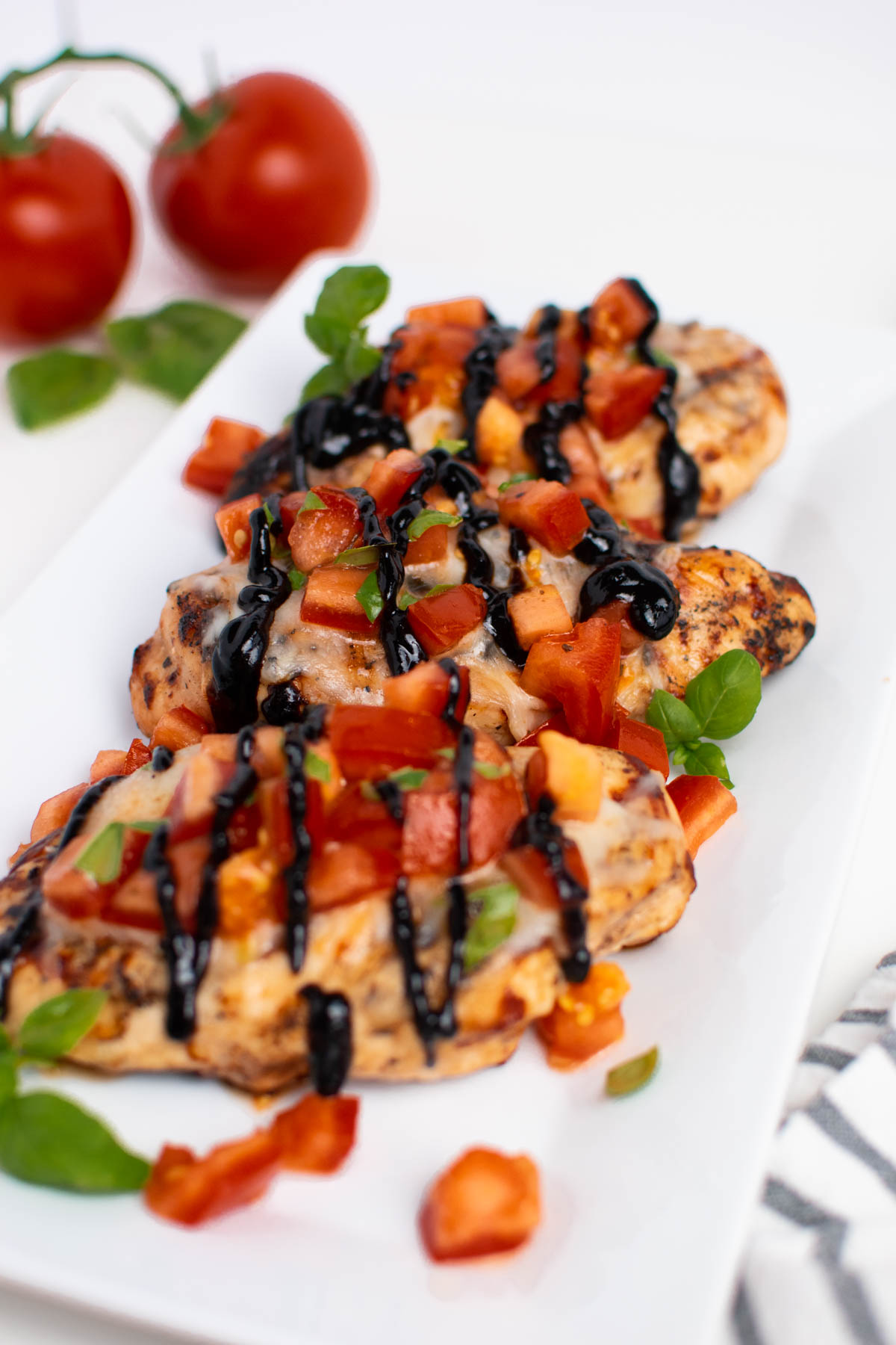 Three pieces of grilled bruschetta chicken on white platter with fresh tomatoes in background.
