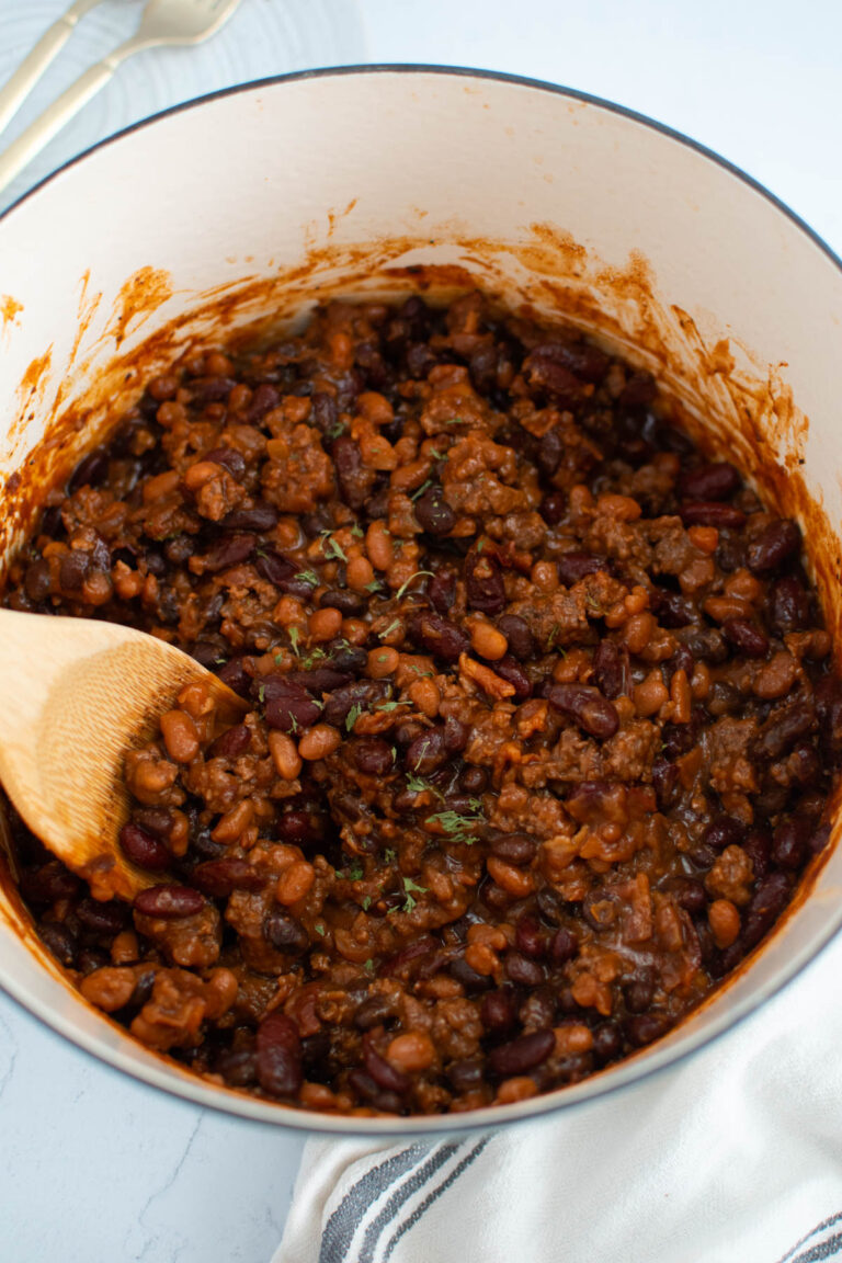 Cowboy baked beans in dutch oven with wooden spoon and striped towel nearby.