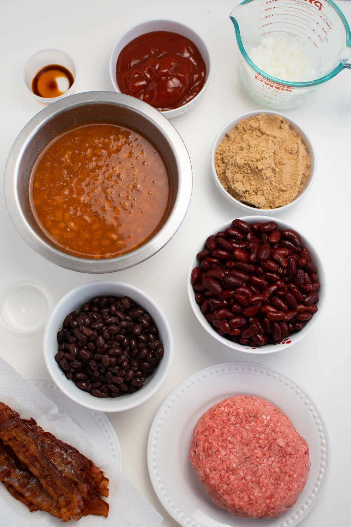 Bowls of ingredients for cowboy baked beans including kidney beans, pork and beans, and bacon.