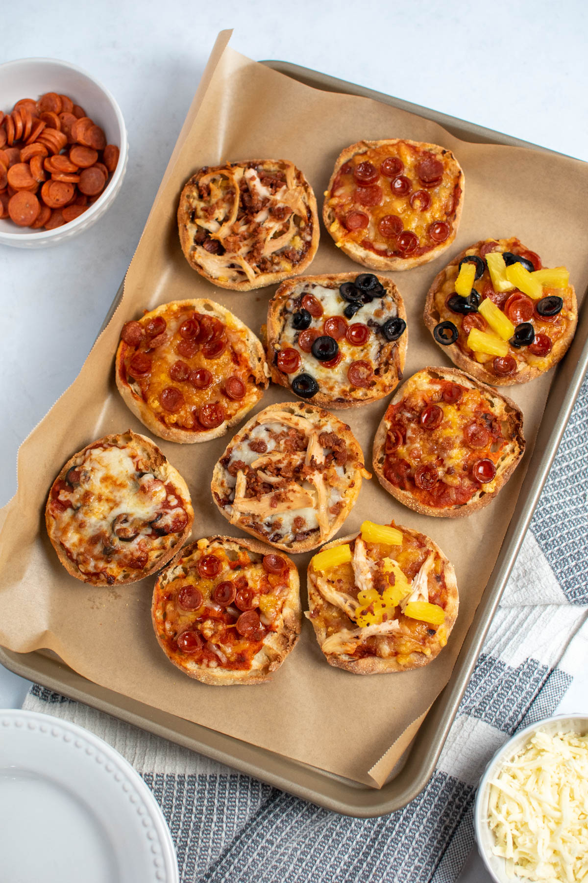 Several English muffin mini pizzas on parchment paper lined baking sheet next to kitchen towel.