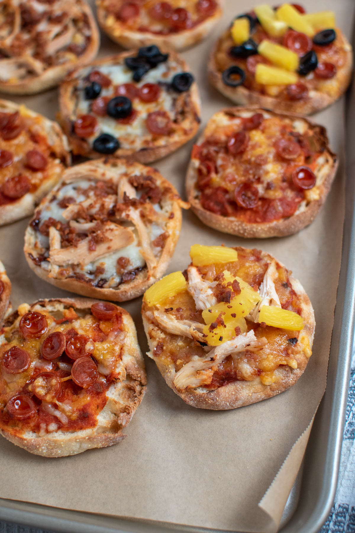 English muffin pizzas with pepperoni, pineapple, and chicken on baking sheet.