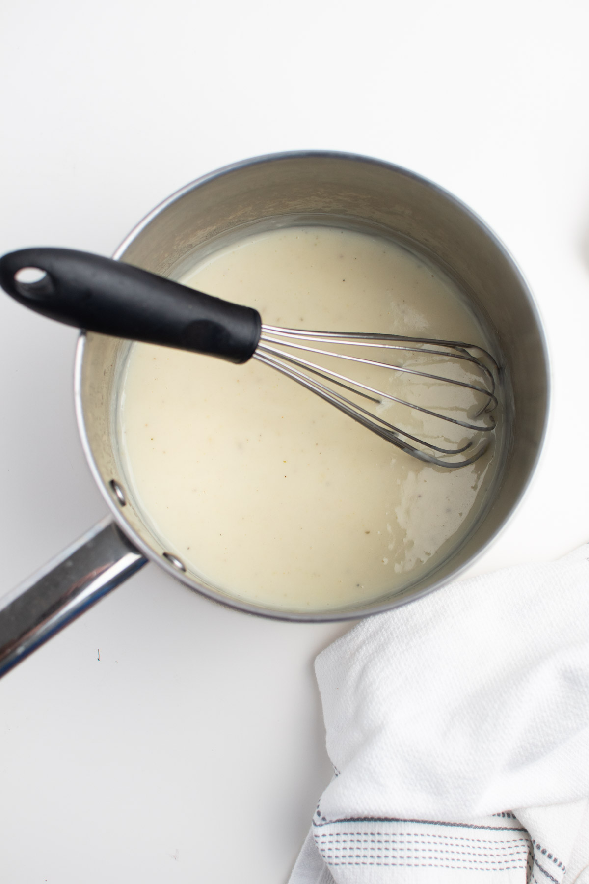White gravy in pot metal sauce pot with wire whisk next to white kitchen towel.