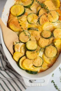 Zucchini and squash casserole in white baking dish with wood spoon and sprigs of thyme.