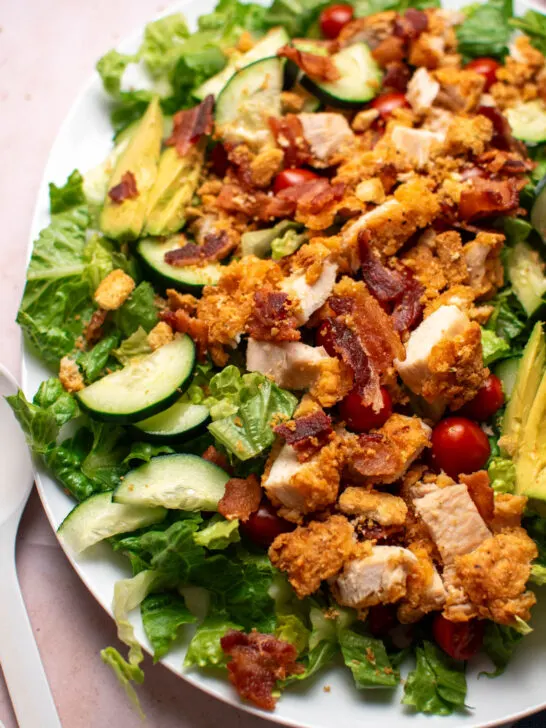 BLT salad with cucumbers, crispy chicken, and avocado on white platter.