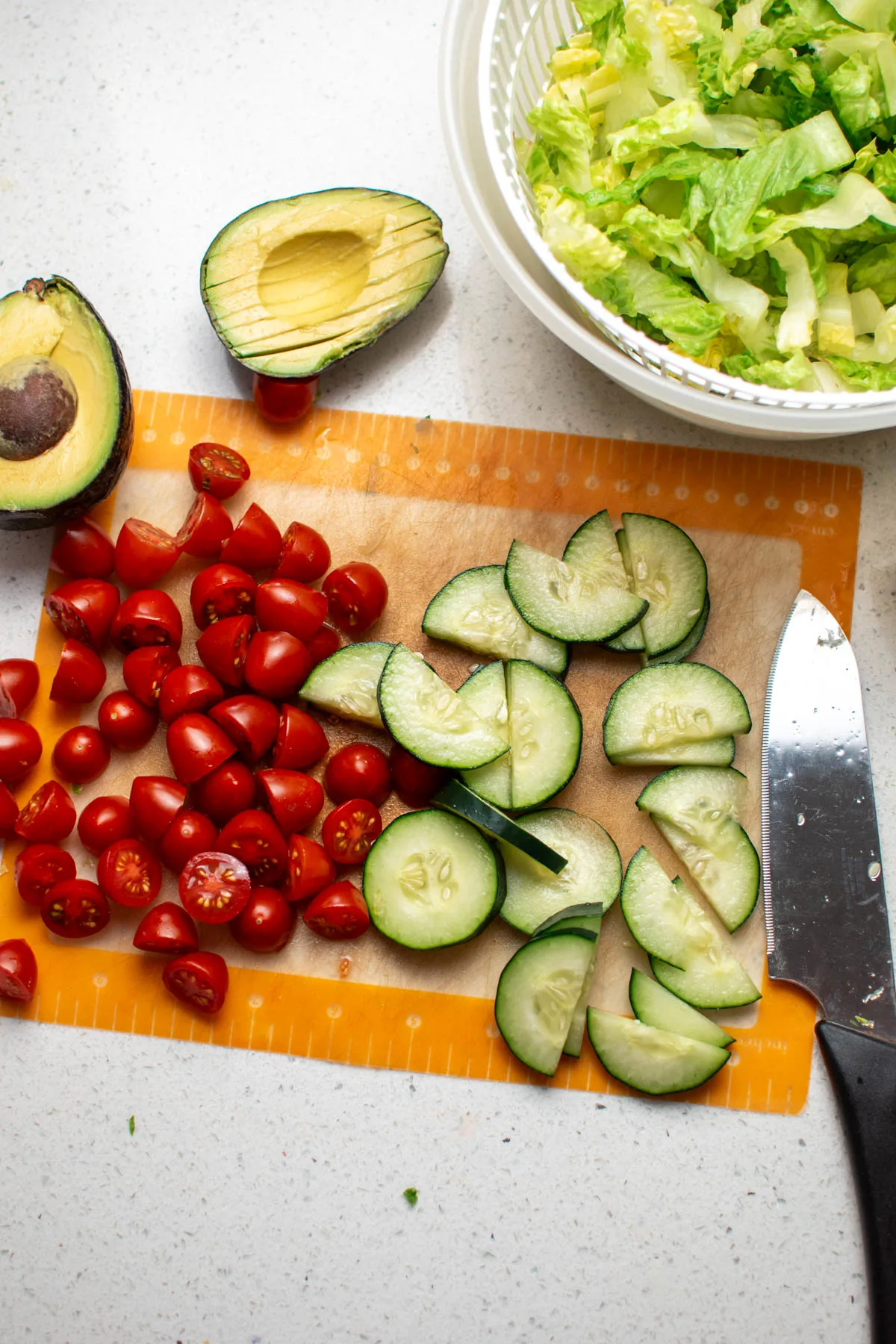 Chopped tomatoes, cucumbers, and avocado halves next to bowl of washed lettuce.