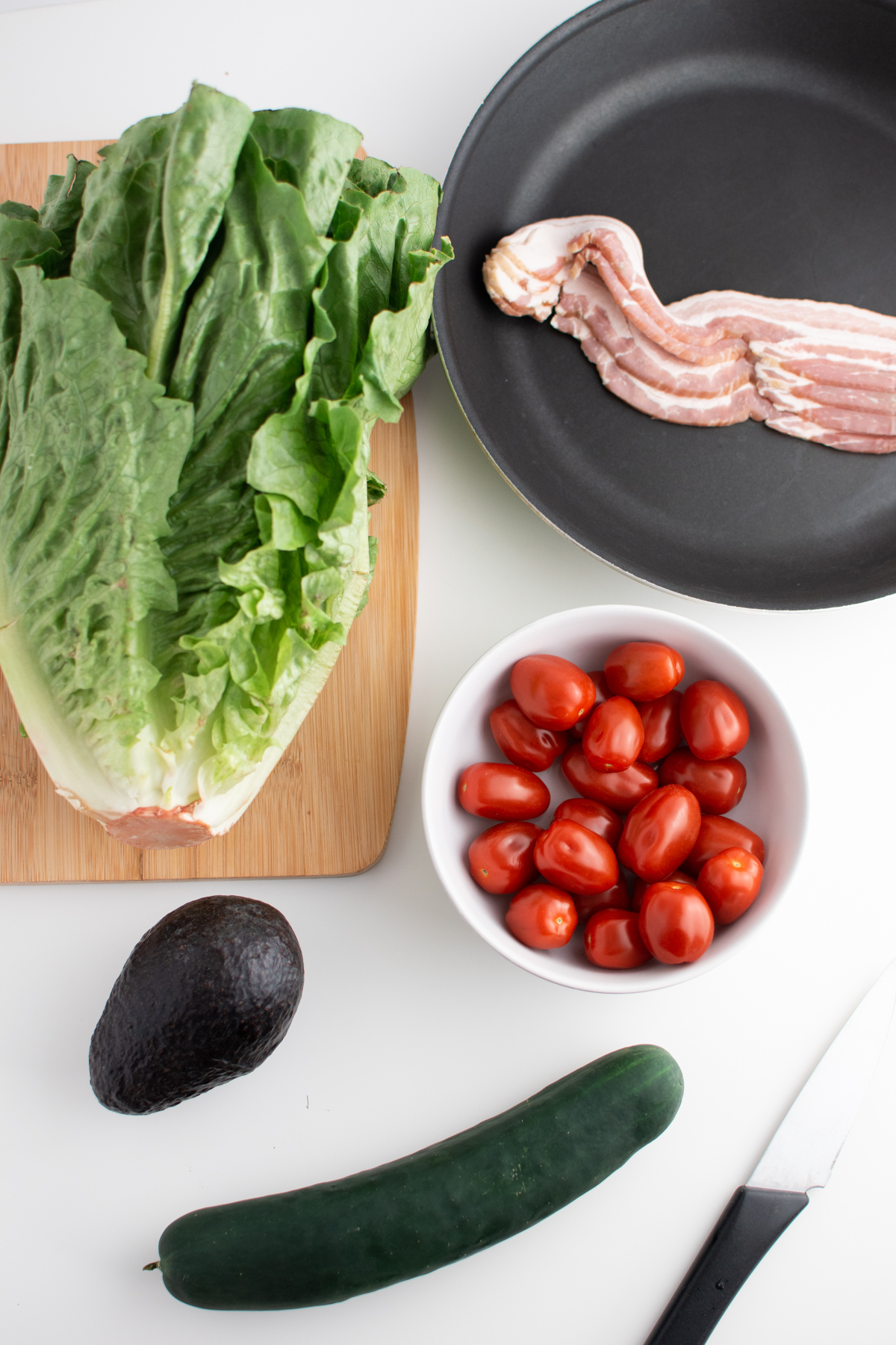 BLT salad ingredients including uncooked bacon, cherry tomatoes, avocado, cucumber and lettuce.