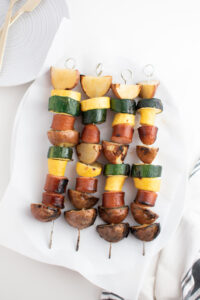 Four grilled sausage kabobs with squash and potatoes on parchment paper and white platter.