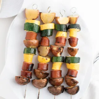 Four grilled sausage kabobs with squash and potatoes on parchment paper and white platter.