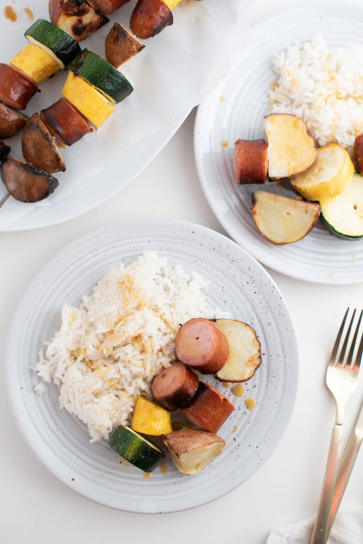 Two plates of grilled sausage, potatoes, squash, and rice on white table next to gold utensils.