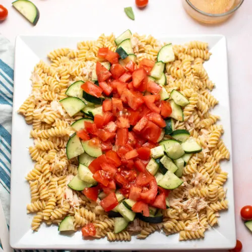 Rotini pasta salad with tomatoes, cucumber, and chicken on large white platter with ingredients scattered around.