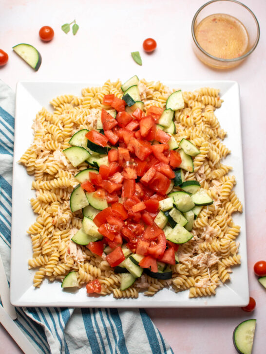 Rotini pasta salad with tomatoes, cucumber, and chicken on large white platter with ingredients scattered around.