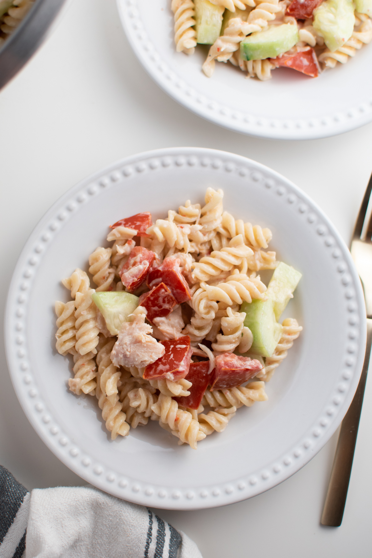 Rotini pasta salad on white dinner plates with gold fork and kitchen towel.