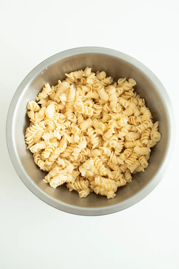 Plain, cooked rotini pasta in a large metal mixing bowl with white background.