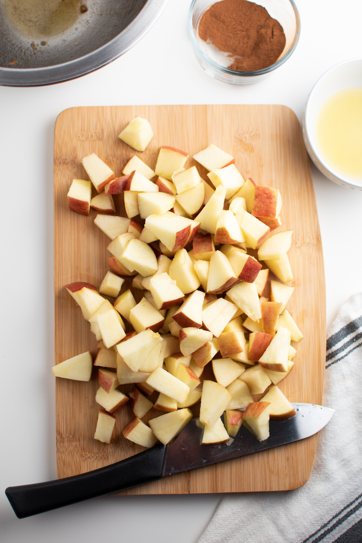 Chopped apples on wood cutting board surrounded by knife and bowls of ingredients.