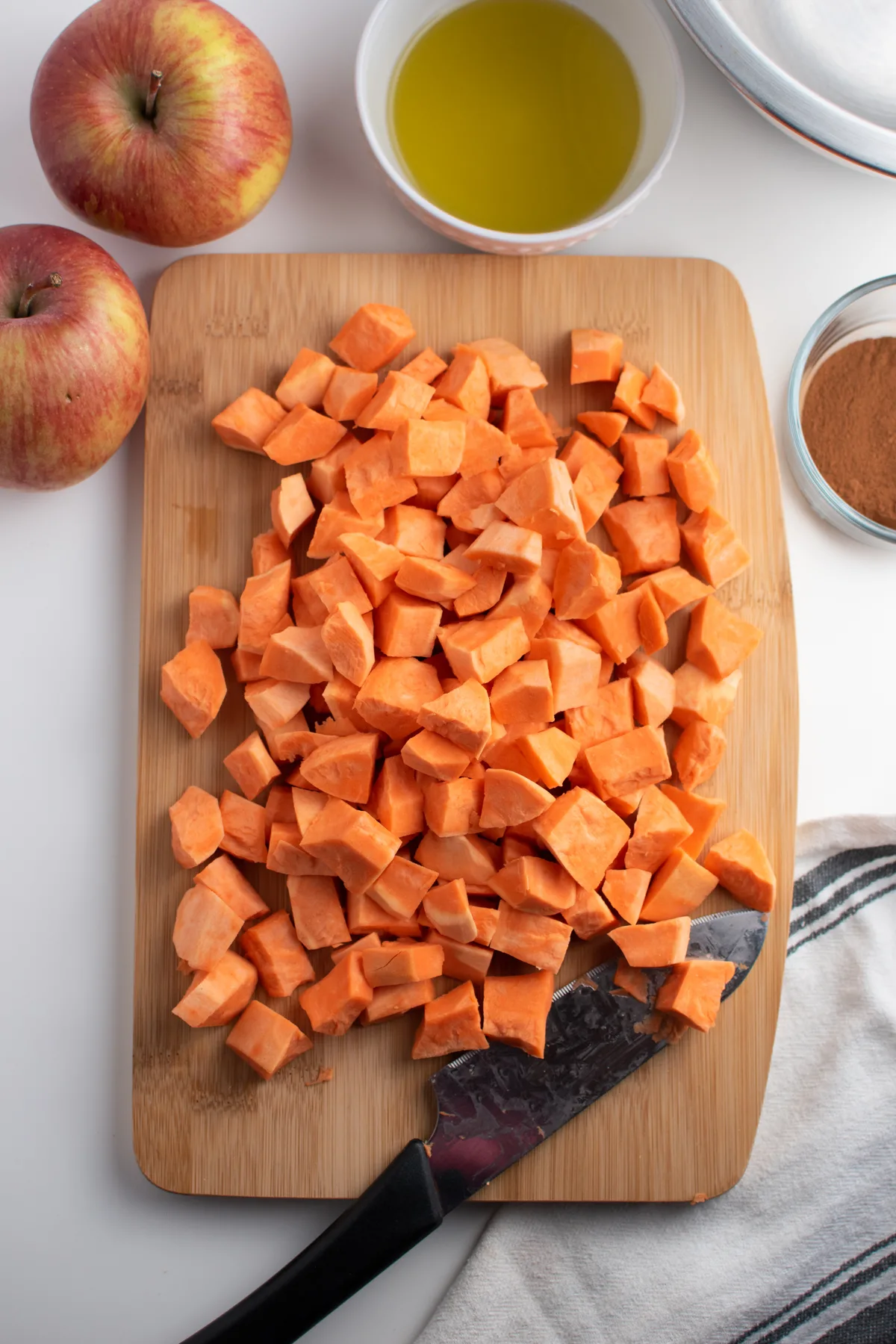 Chopped sweet potato cubes on a cutting board surrounded by knife, apples and bowls of ingredients.