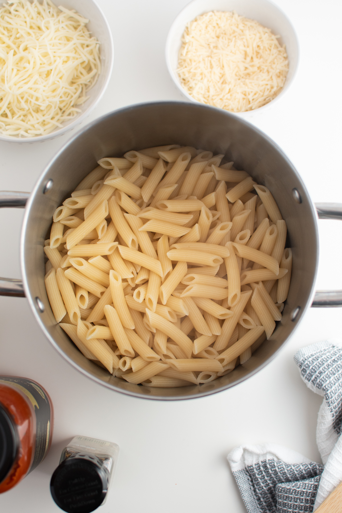Cooked penne pasta in large metal stock pot next to cheeses and sauce.