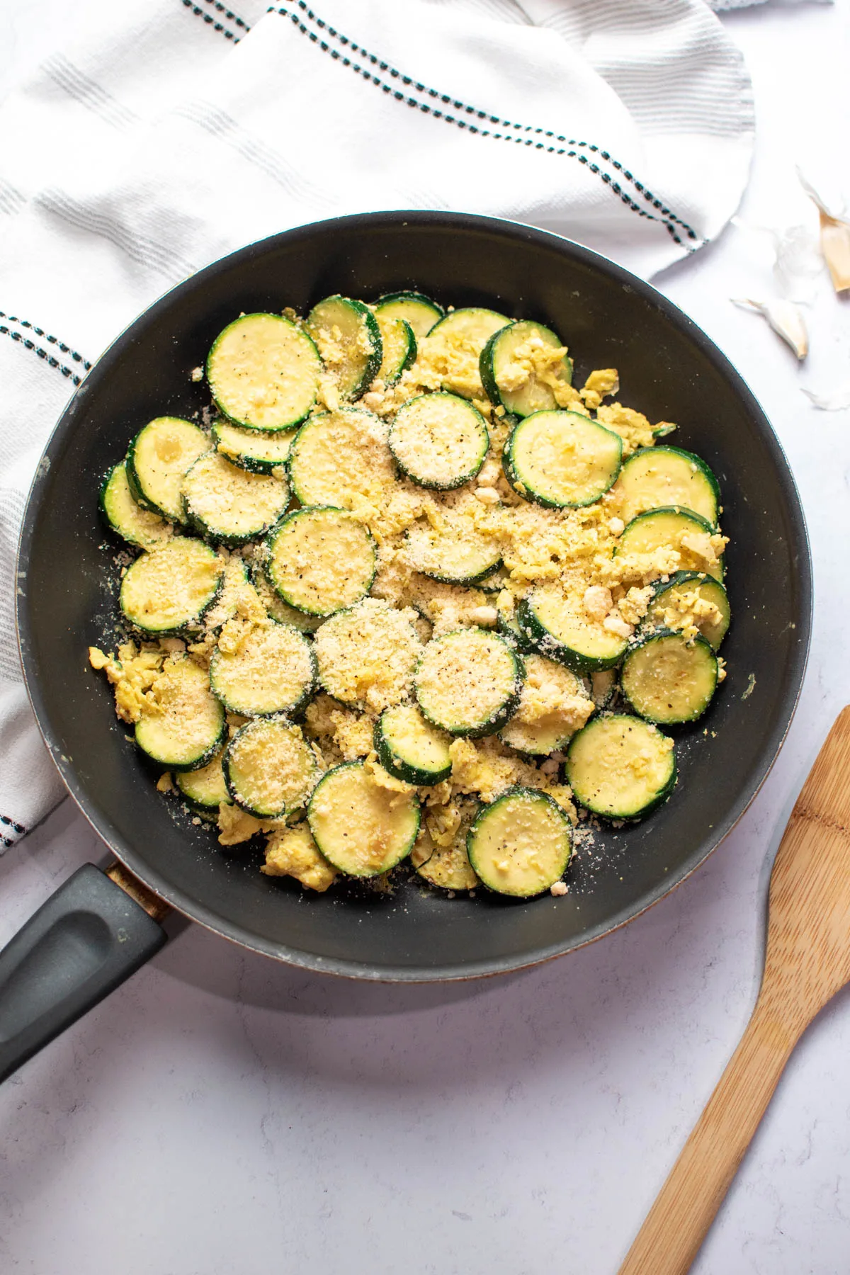 Cooked eggs and zucchini with parmesan cheese in black frying pan with wood spoon laying nearby.