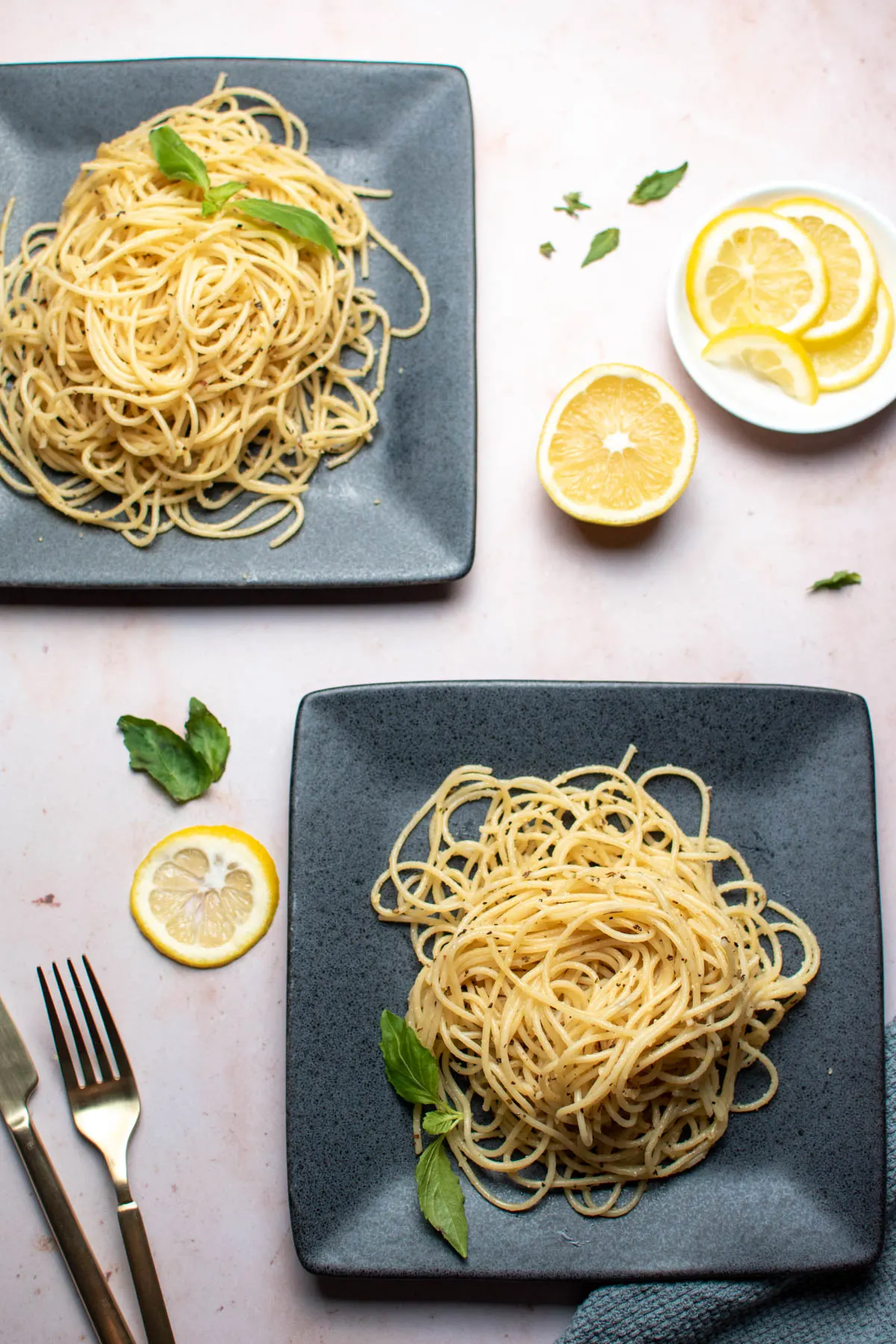 Two gray square plates with lemon and basil pasta, fresh lemon slices, and gold utensils on countertop.