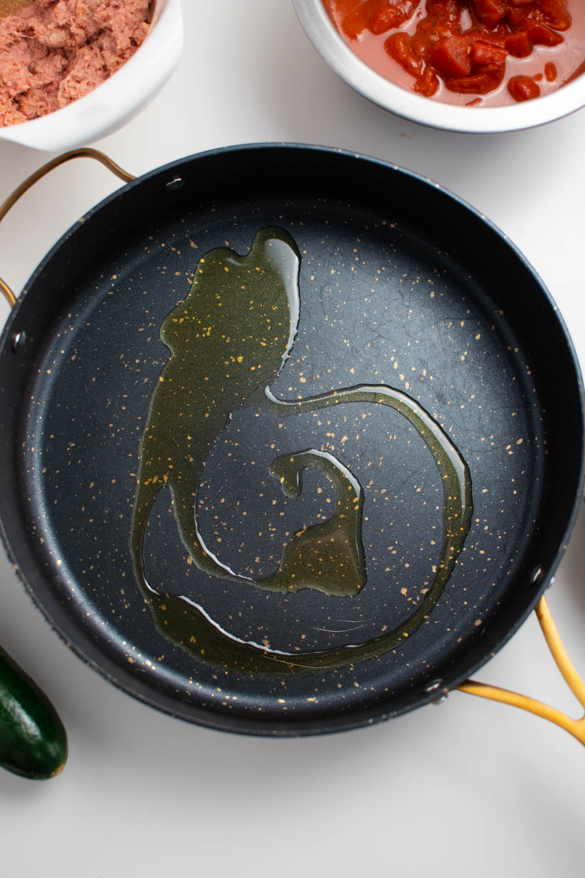 Swirl of olive oil in large black speckled skillet, with bowls of other ingredients nearby.