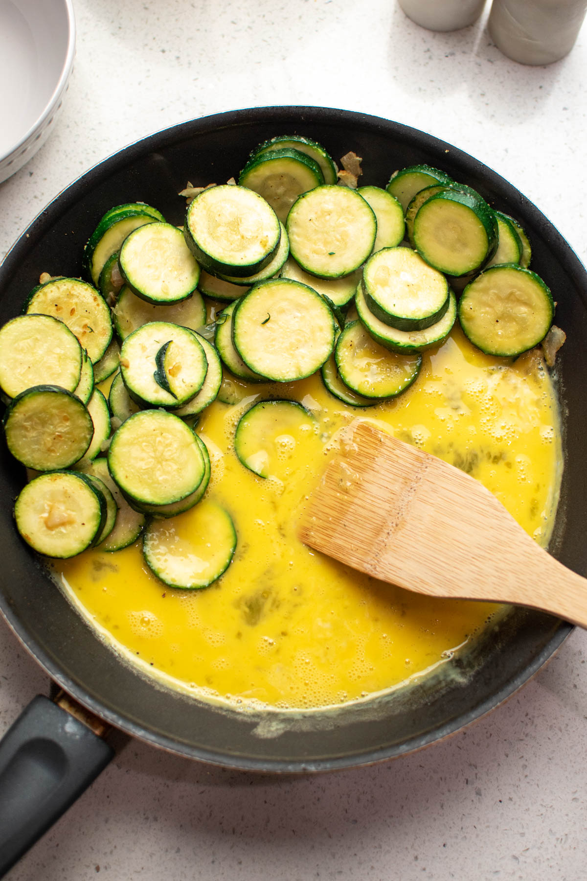 Black frying pan with raw eggs, sauteed zucchini, and wood spoon on countertop.