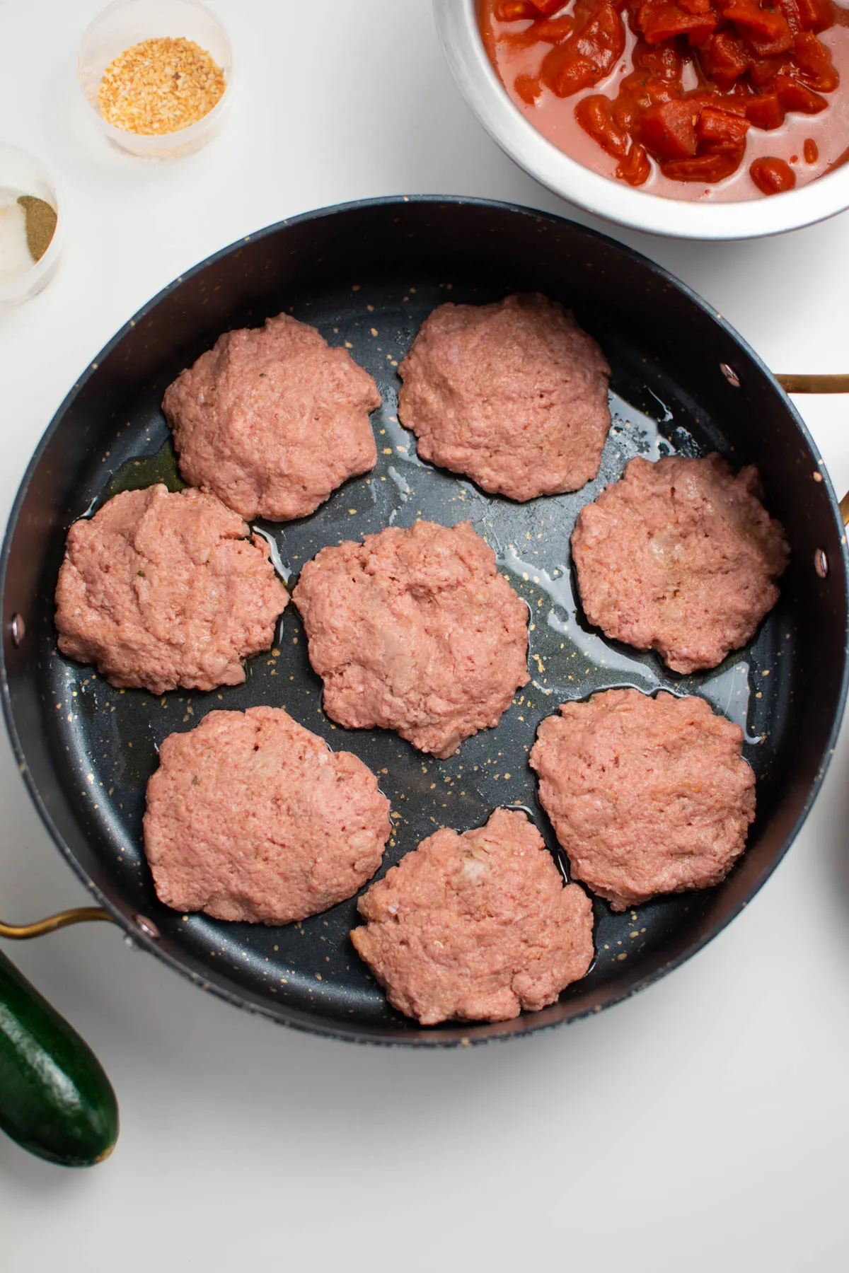 Eight raw turkey patties in large black skillet with bowl of diced tomatoes nearby.