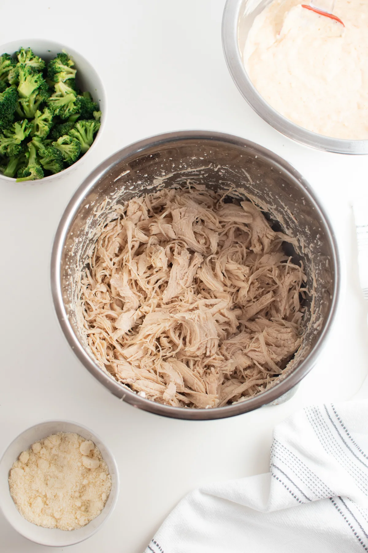Shredded chicken in large metal mixing bowl surrounded ingredients in prep bowls.