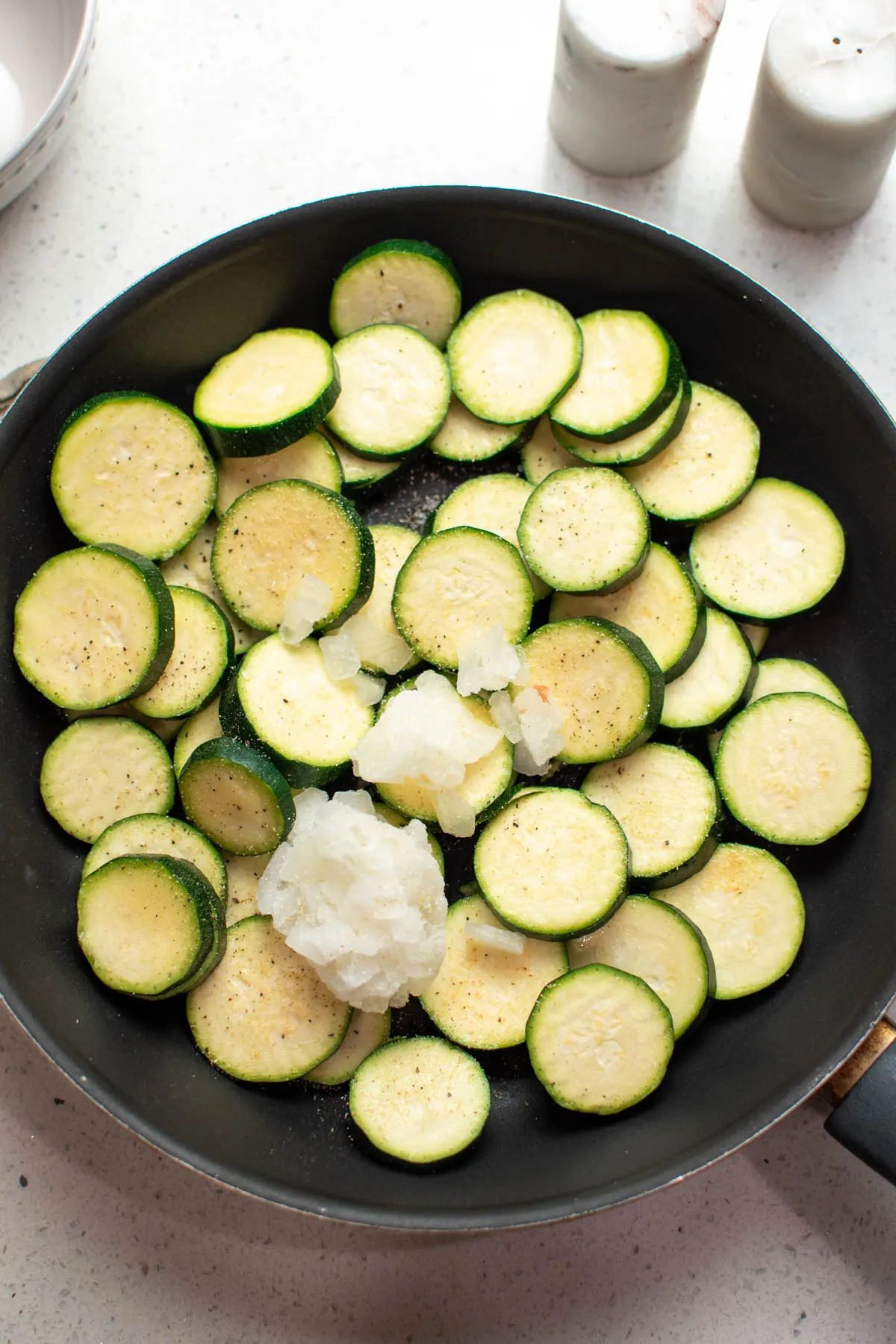 Black frying pan with raw sliced zucchini, frozen onions, and seasoning on quartz countertop.