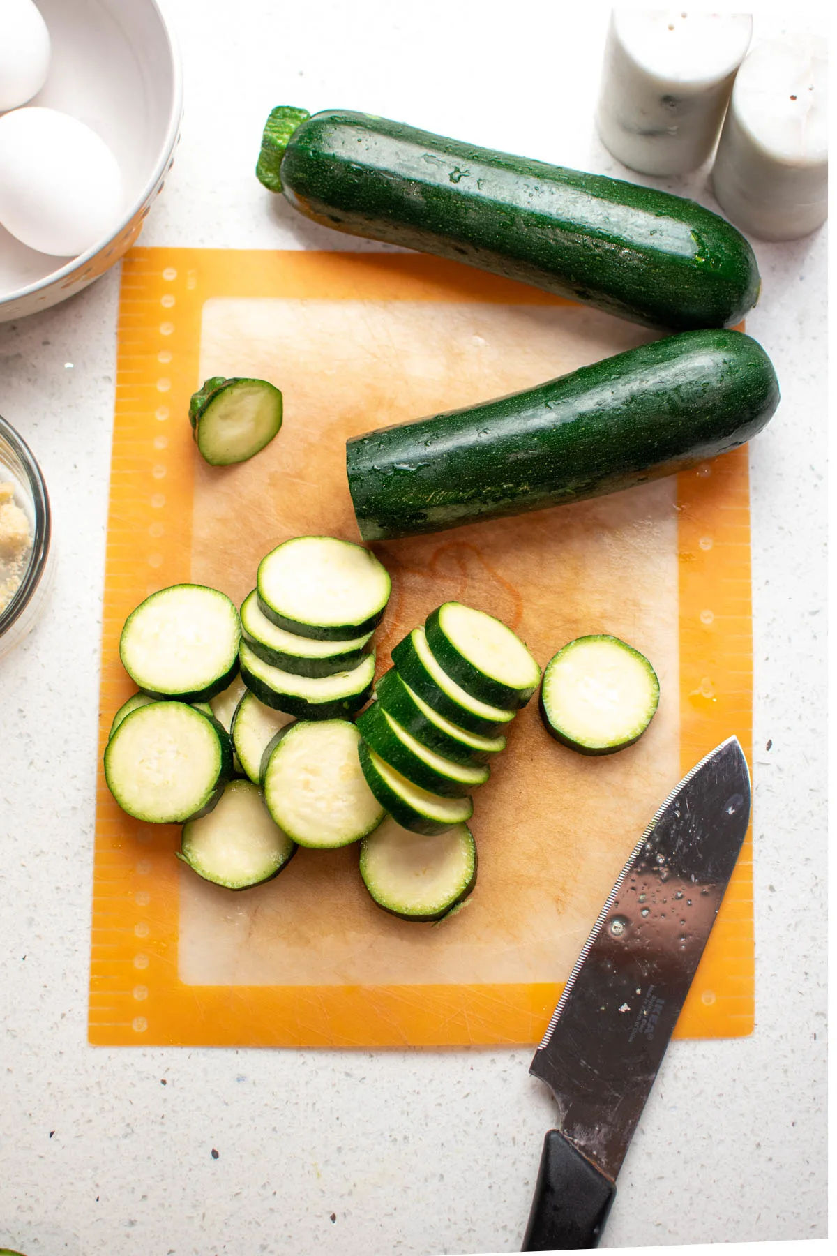 Countertop with partially sliced zucchini and a knife resting on orange cutting board.