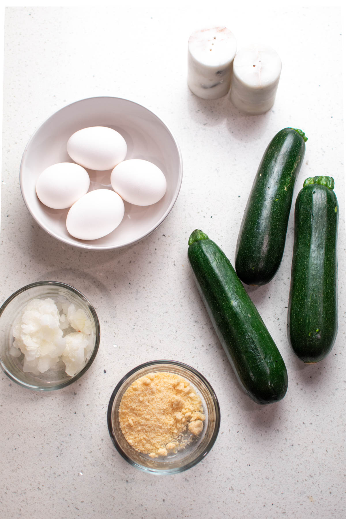 Ingredients for zucchini scrambled eggs on quartz countertop including eggs, onion, and parmesan cheese.