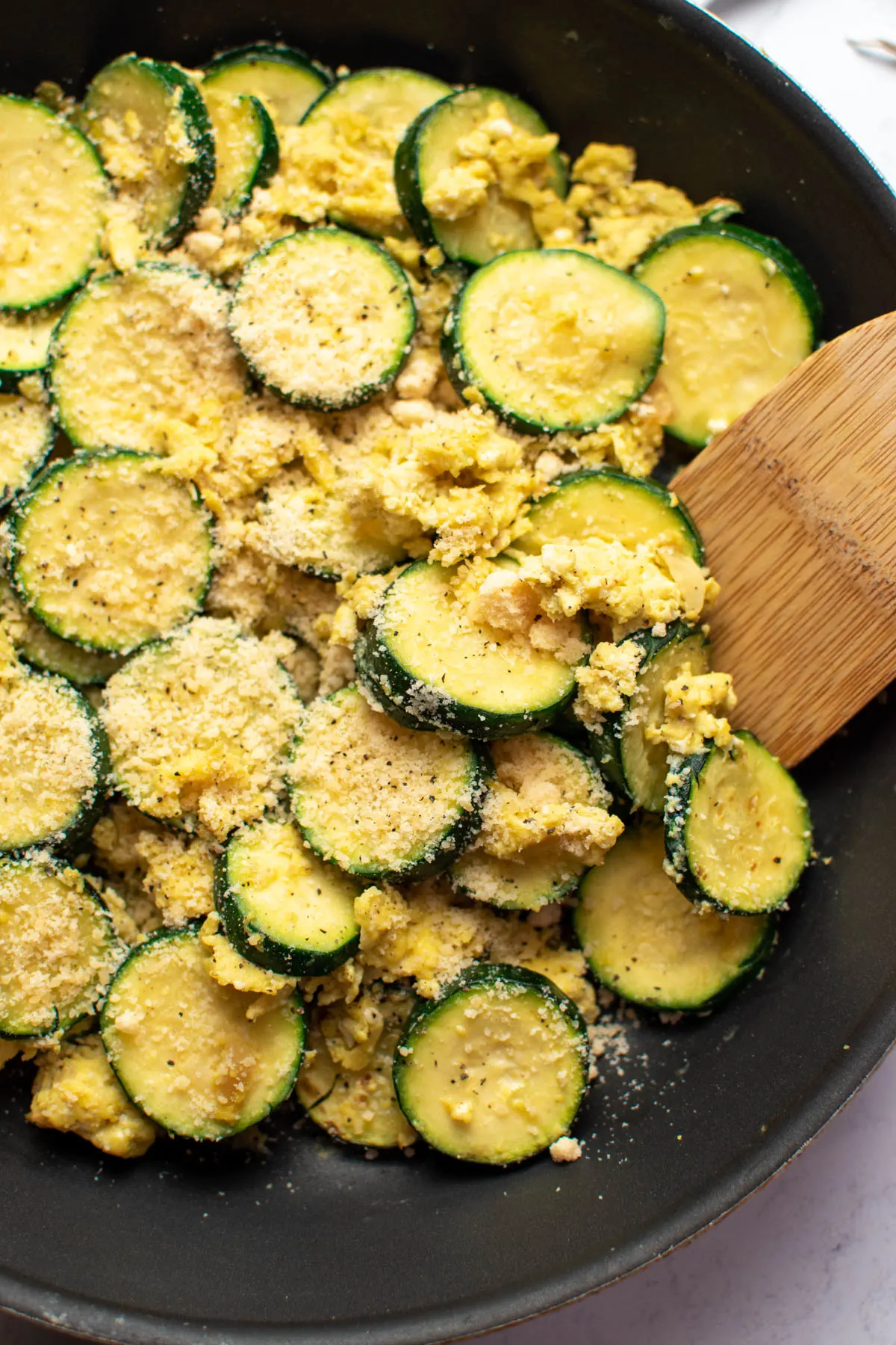 Wood spoon rests in black frying pan with zucchini scrambled eggs and parmesan cheese.