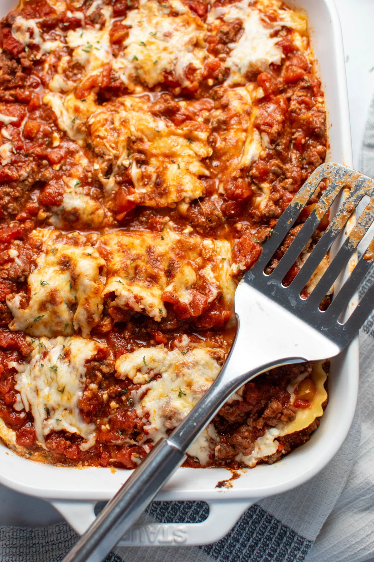 Metal spatula rests on white baking dish with cut pieces of lasagna with melted mozzarella.