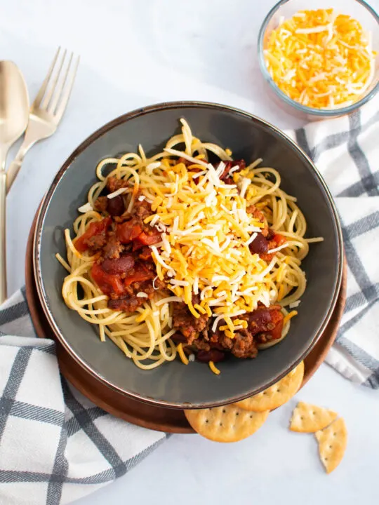 Bowl of chili spaghetti with shredded cheese surrounded by checkered towel, silverware, bowl of cheese, and crackers.