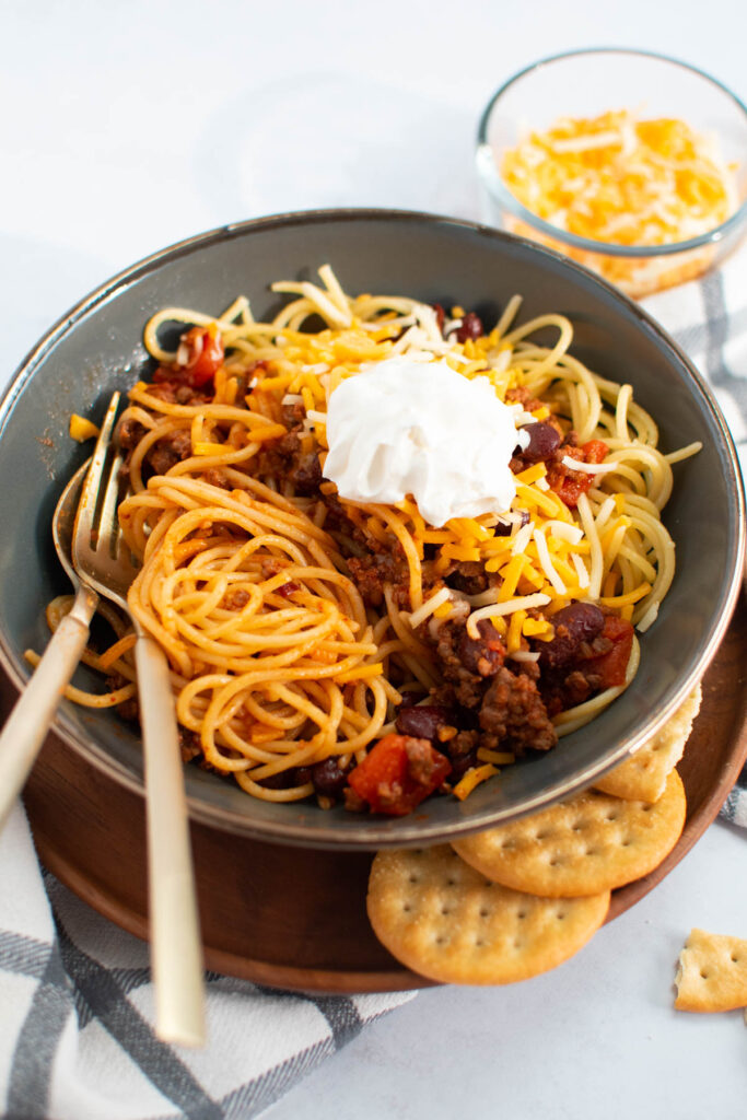 Fork and spoon in green bowl of Cincinatti chili topped with cheese and sour cream.