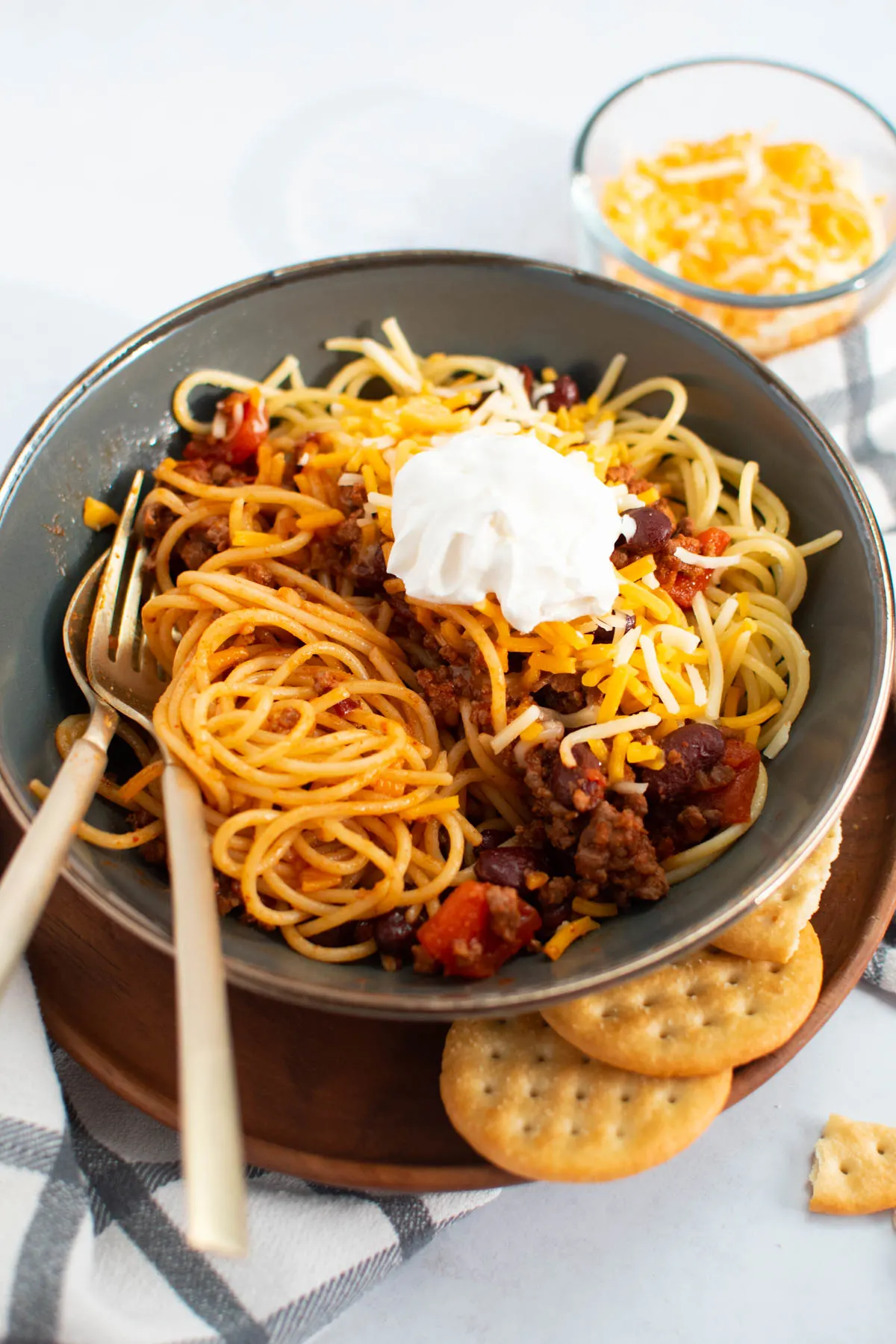 Fork and spoon in green bowl of Cincinatti chili topped with cheese and sour cream.
