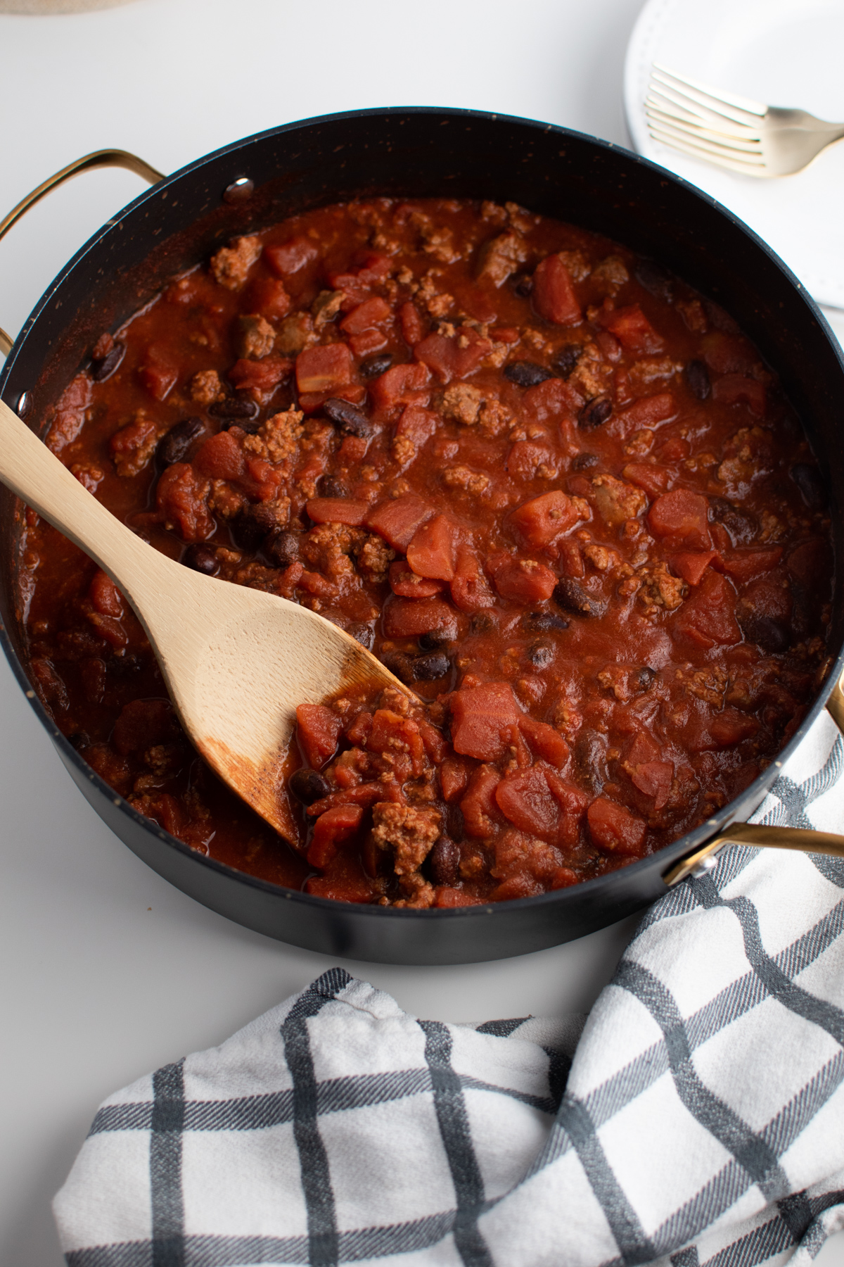 Wooden spoon rests in Cincinnati chili with tomatoes, ground beef, and kidney beans, all in a large skillet. 