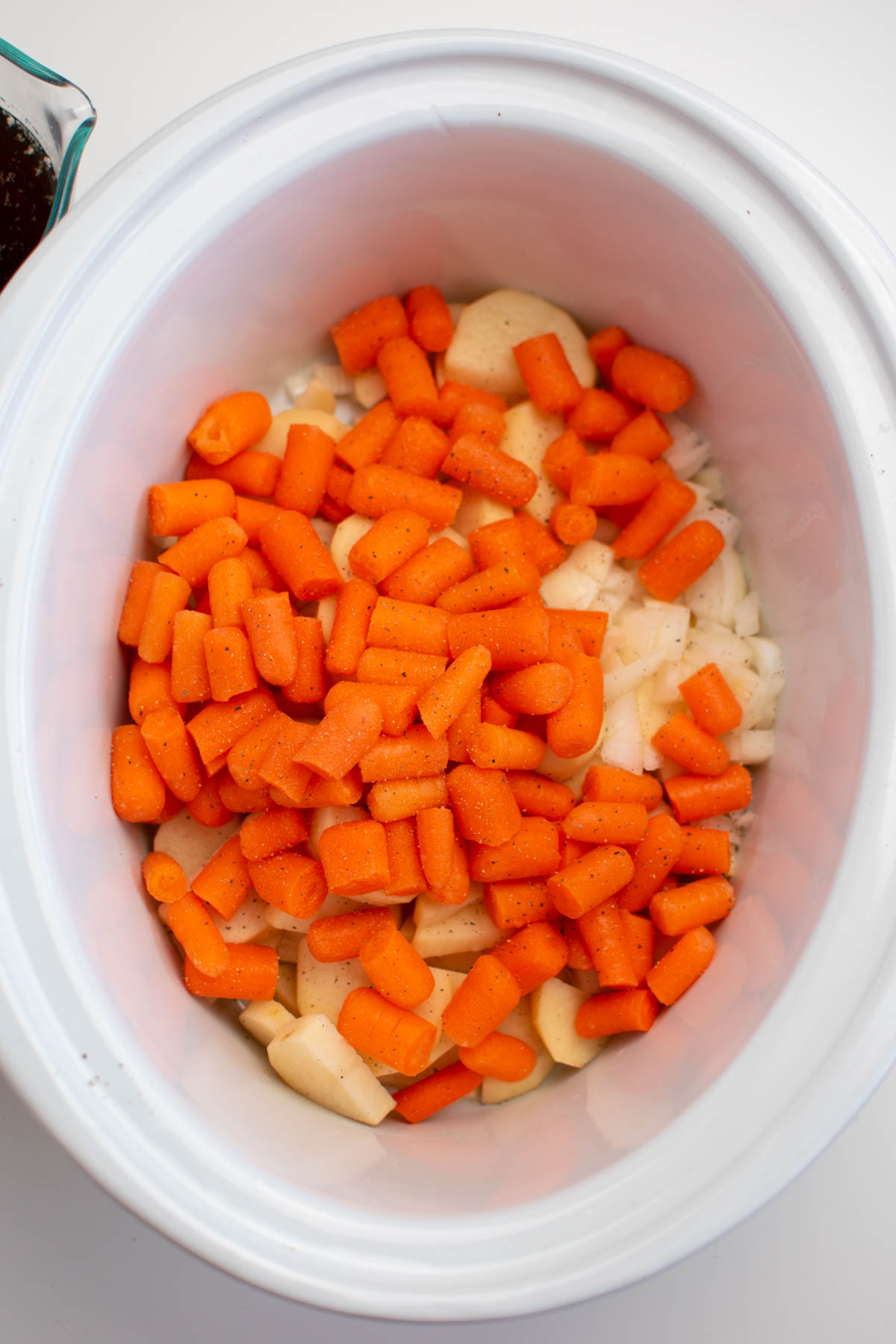 Pieces of raw baby carrots, potatoes, and onion in white Crock Pot insert.