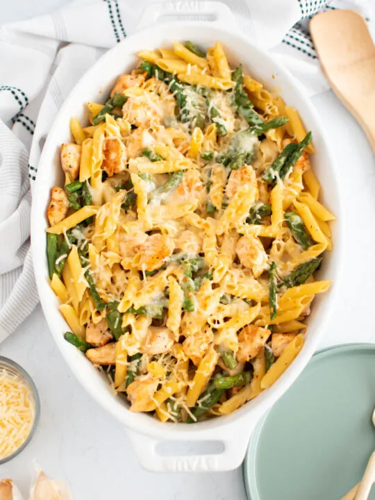 Chicken and asparagus pasta in white baking dish surrounded by plates, towel, and wood spoon.
