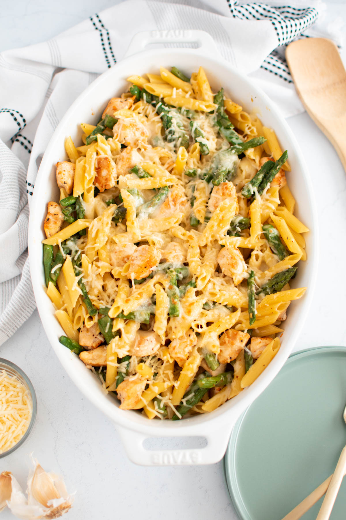 Chicken and asparagus pasta in white baking dish surrounded by plates, towel, and wood spoon.