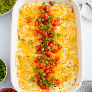 Pan of creamy chicken enchiladas with tomato and green onion garnish on table with bowls of toppings nearby.