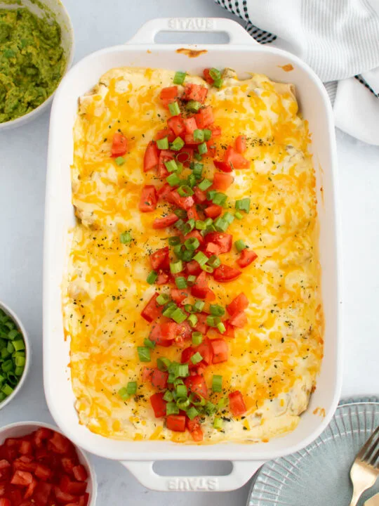 Pan of creamy chicken enchiladas with tomato and green onion garnish on table with bowls of toppings nearby.