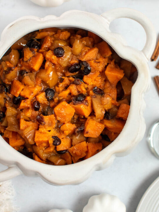 Slow cooker sweet potatoes and apples in white pumpkin dish with white pumpkin decor nearby.