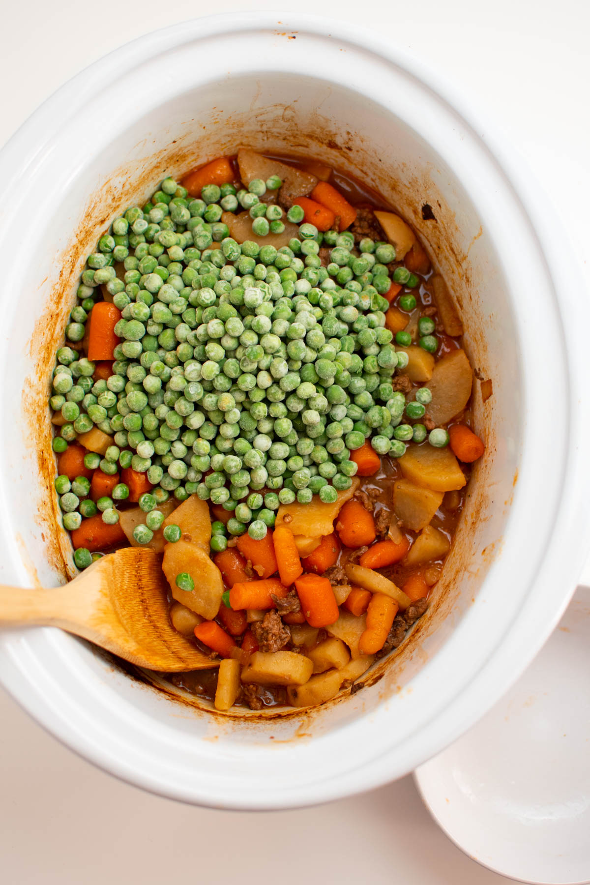 Pile of frozen peas on top of cooked hamburger stew in white Crock Pot insert.