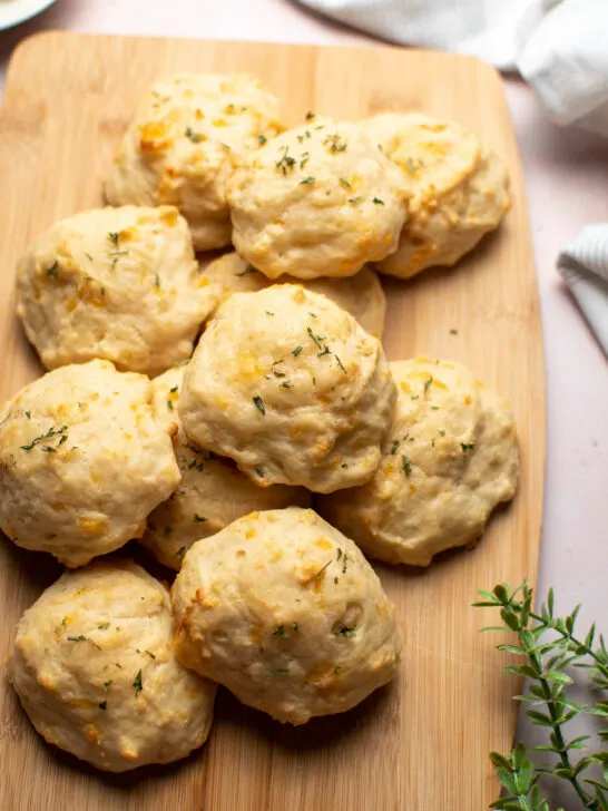 Parmesan drop biscuits arranged on wood cutting board with greenery stem nearby.