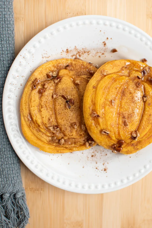 Pumpkin shaped pancakes covered in pecans, syrup, and cinnamon on white plate.