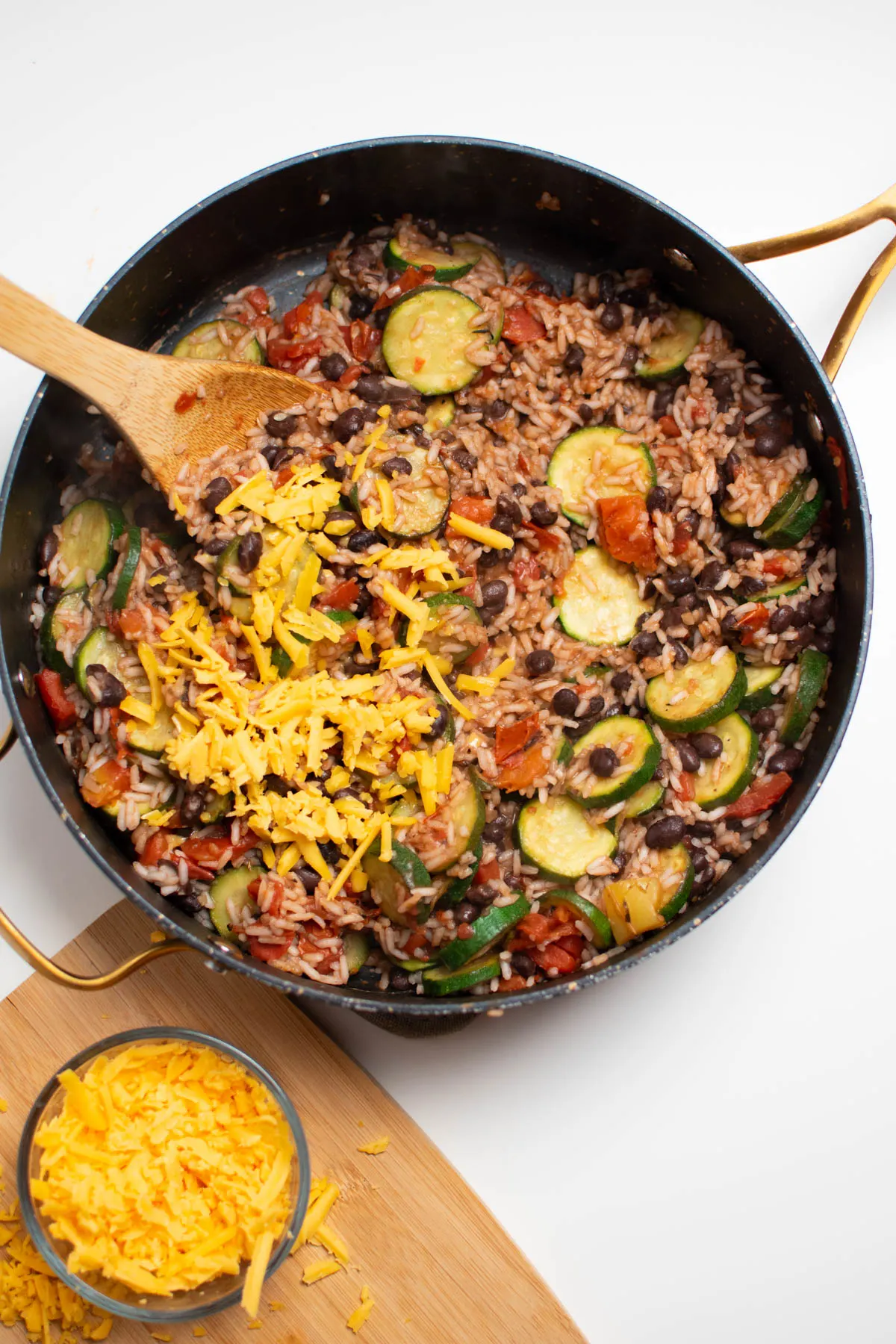A small amount of shredded cheddar cheese on black skillet full of zucchini, rice, and black beans.