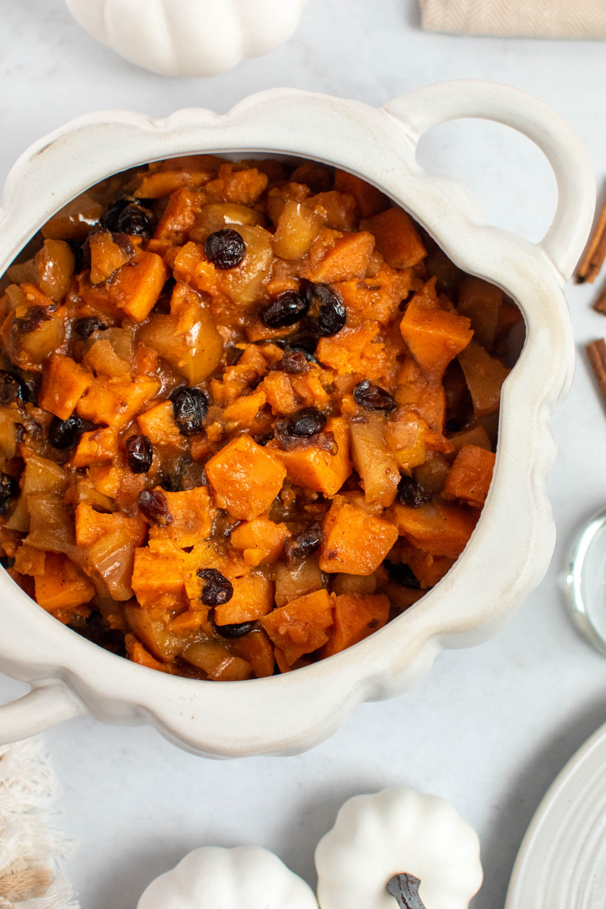 Slow cooker sweet potatoes and apples in white pumpkin dish with white pumpkin decor nearby.