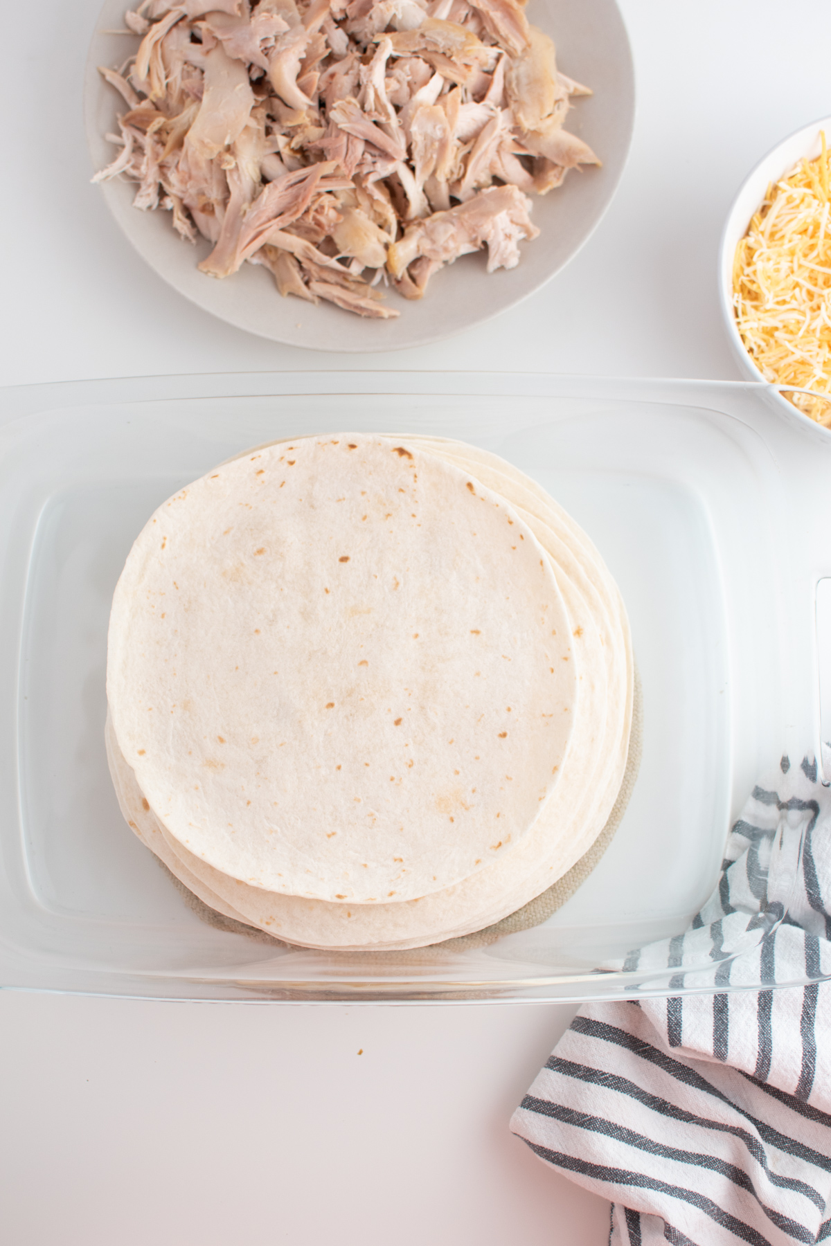 Large stack of warmed tortillas in glass pan next to prep bowls with cheese and chicken.