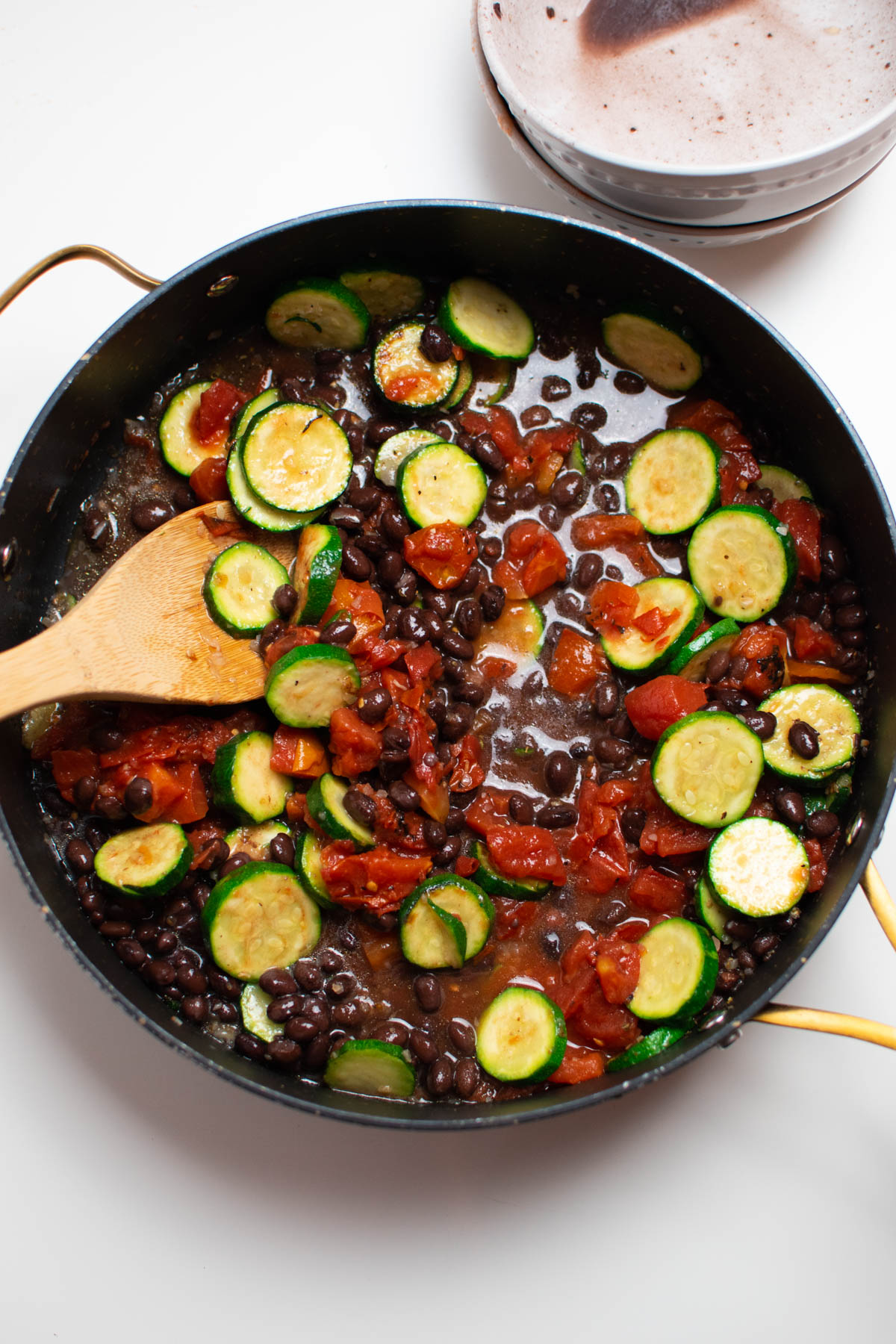 Zucchini, tomatoes, and black beans in large black skillet with wooded spoon.