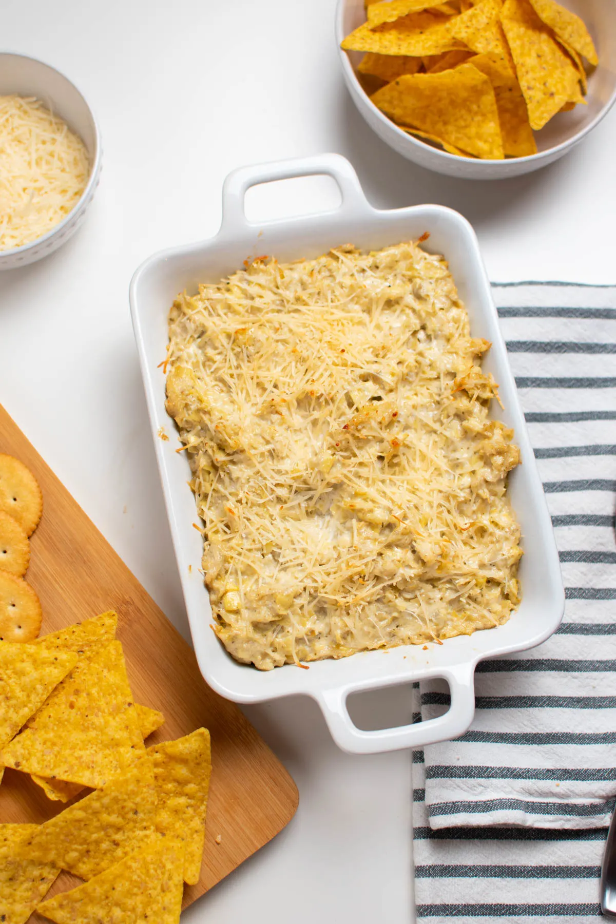 Artichoke parmesan dip in small white casserole baking dish with chips and cheese.